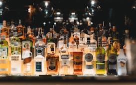 A collection of assorted liquor bottles is arranged on a backlit shelf. The bottles vary in shape, size, and color, featuring well-known brands like Absolut, Bacardi, Martini, and J&auml;germeister. The background is dimly lit, creating a reflective and ambient setting.