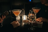 A dimly lit setting with two cocktails in martini glasses placed on a round table. Two people are partially visible, each with manicured hands. A lit candle in a glass holder adds a warm glow. There is also a glass of white wine, a smartphone, and a small bowl with snacks. A package of smoking accessories is placed on the table, hinting at a relaxed, social gathering.