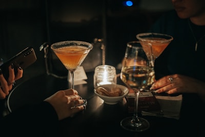 A dimly lit setting with two cocktails in martini glasses placed on a round table. Two people are partially visible, each with manicured hands. A lit candle in a glass holder adds a warm glow. There is also a glass of white wine, a smartphone, and a small bowl with snacks. A package of smoking accessories is placed on the table, hinting at a relaxed, social gathering.