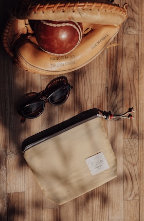 A baseball glove and ball made of handcrafted top grain cowhide lie on a wooden surface. Nearby, a pair of dark sunglasses and a beige zipper pouch with a small tag are placed. The lighting casts soft shadows, enhancing the warm tones of the objects.