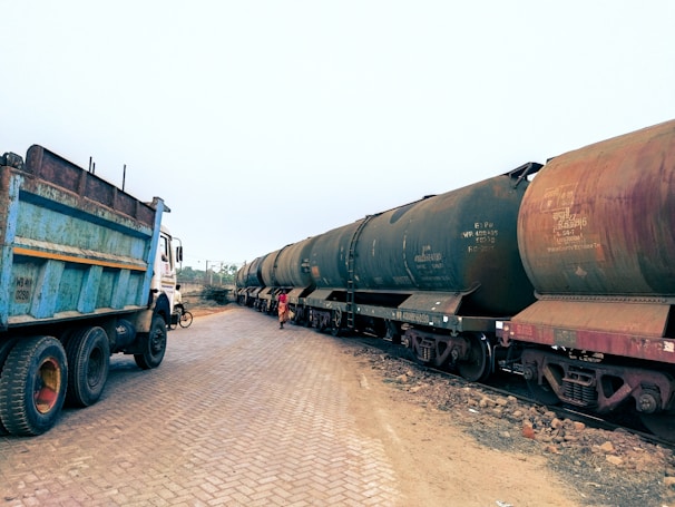An outdoor scene showing a large blue industrial truck parked on a brick-paved road near several rust-colored train tank wagons. A person is walking beside the train wagons, with a few scattered rocks seen on the ground.