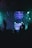 A dark environment with dim lighting and silhouettes of people. Bright teal and purple lights create a vibrant atmosphere. Particles or confetti fill the air, catching the colorful light.