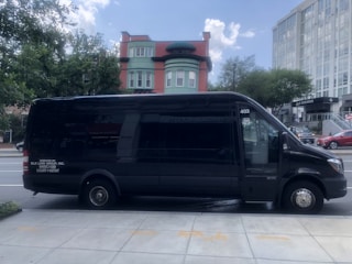 A dark-colored minibus is parked on the side of an urban street. In the background, there are buildings with various architectural styles, including a red and green building with distinct windows and a modern glass structure. Trees provide greenery on either side of the street.