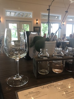 A wine tasting setup on a wooden table, with a large empty wine glass, a holder containing three small bottles with different colored liquids, and a potted plant with thin branches and a white flower. The background shows a bright room with large windows and several people sitting at tables.