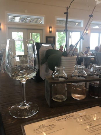 A wine tasting setup on a wooden table, with a large empty wine glass, a holder containing three small bottles with different colored liquids, and a potted plant with thin branches and a white flower. The background shows a bright room with large windows and several people sitting at tables.