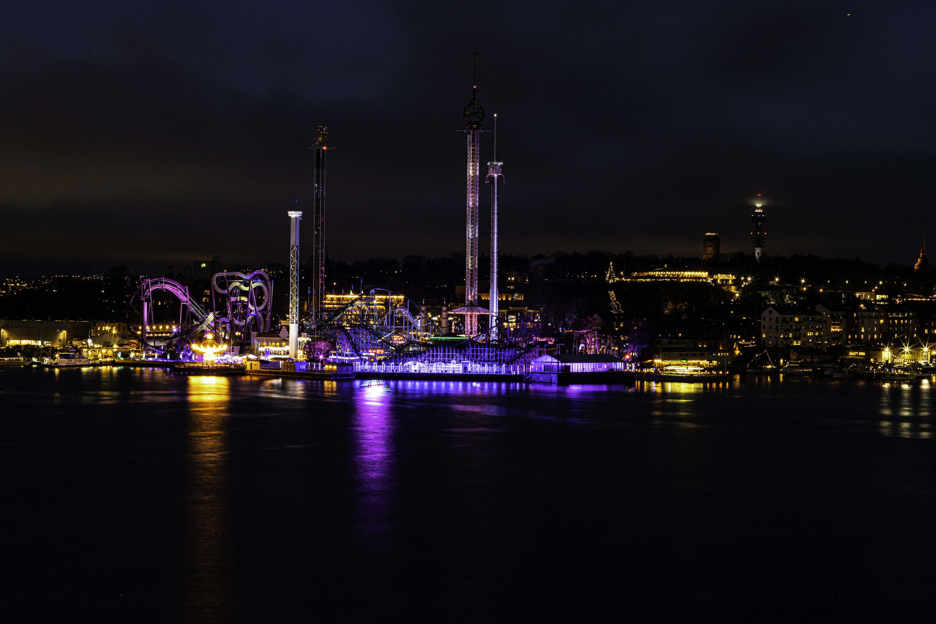 An illuminated waterfront amusement park glows purple and gold, reflected in dark water at night.