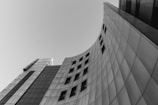 A modern building with a curved, sleek architectural design, featuring a series of rectangular windows. The facade is composed of panels, reflecting a minimalist and contemporary style.