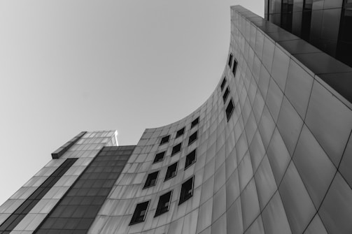 A modern building with a curved, sleek architectural design, featuring a series of rectangular windows. The facade is composed of panels, reflecting a minimalist and contemporary style.