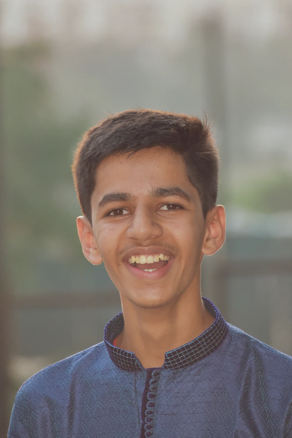 The image shows a young boy with short brown hair wearing a blue and white patterned shirt, smiling brightly at the camera. The background is blurred, suggesting an outdoor setting or a busy street scene.