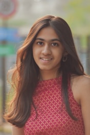 A young woman with long, dark hair is smiling directly at the camera. She is wearing a sleeveless red dress with a subtle pattern and round earrings. The background is slightly blurred, showing some outdoor elements like trees, a street sign, and a fence.