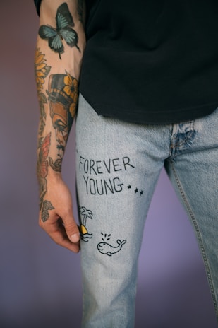 A person's arm with colorful tattoos featuring flowers, a butterfly, and various designs. The arm is resting against light blue denim jeans, which have black text reading 'FOREVER YOUNG' alongside small drawings of a palm tree, sun, and whale.