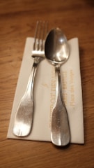 A silver fork and spoon are placed on top of a folded, cream-colored napkin. The napkin rests on a wooden table, and part of the napkin has some text printed on it.