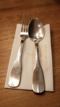 A silver fork and spoon are placed on top of a folded, cream-colored napkin. The napkin rests on a wooden table, and part of the napkin has some text printed on it.