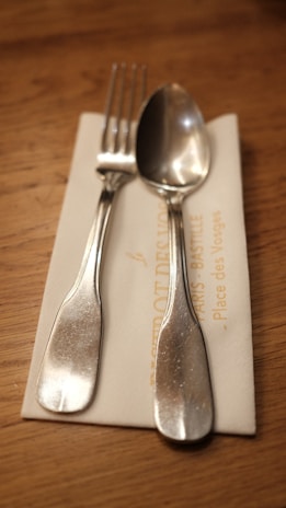 A silver fork and spoon are placed on top of a folded, cream-colored napkin. The napkin rests on a wooden table, and part of the napkin has some text printed on it.