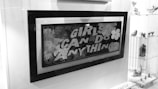 Black and white image showing a framed artwork or poster with the text 'GIRLS CAN DO ANYTHING'. The frame is positioned inside a display case or a storefront. To the right of the frame, there are glass shelves displaying various small figurines and art pieces. The background and surroundings are well-lit.