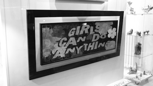 Black and white image showing a framed artwork or poster with the text 'GIRLS CAN DO ANYTHING'. The frame is positioned inside a display case or a storefront. To the right of the frame, there are glass shelves displaying various small figurines and art pieces. The background and surroundings are well-lit.