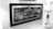 Black and white image showing a framed artwork or poster with the text 'GIRLS CAN DO ANYTHING'. The frame is positioned inside a display case or a storefront. To the right of the frame, there are glass shelves displaying various small figurines and art pieces. The background and surroundings are well-lit.