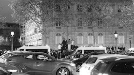 A busy urban scene with a large gathering of people in front of a grand building. Several cars are parked around, and there are trees with sparse foliage. Two street lamps illuminate the area. People are standing on a raised platform, possibly speaking or addressing the crowd.