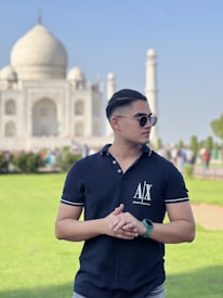 A young man wearing sunglasses and a dark polo shirt stands in front of the Taj Mahal. He has short hair and is clasping his hands. The iconic white marble mausoleum is prominently visible in the background, with clear skies and green grass surrounding the area.