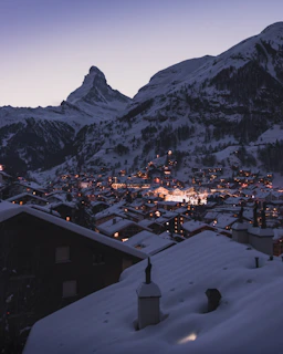 A cozy mountain village covered in snow with warm lights glowing from windows at dusk