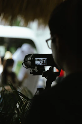 A person is viewed from behind while operating a video camera on a tripod, focusing on a live screen showing another scene in black and white. The setting seems to have natural light, with blurred background elements including greenery and possibly other people.