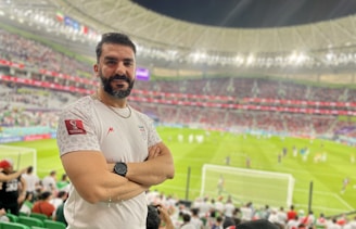 A person stands in a stadium filled with spectators during a sporting event. The grass field and goalposts are visible in the background. The individual is wearing a white jersey with a sports logo and is smiling at the camera.