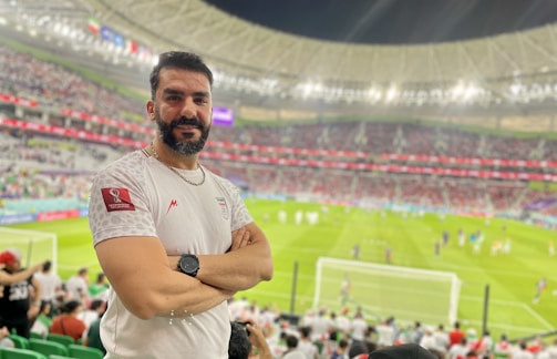 A person stands in a stadium filled with spectators during a sporting event. The grass field and goalposts are visible in the background. The individual is wearing a white jersey with a sports logo and is smiling at the camera.