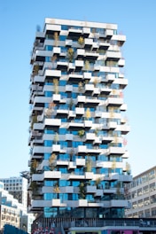 A modern, high-rise building featuring a unique architectural design with staggered balconies adorned with small trees and plants. The structure showcases a combination of blue-tinted glass and white concrete elements, creating a checkerboard pattern. The sky is clear, and surrounding buildings are partially visible.