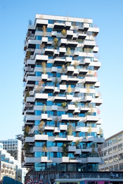 A modern, high-rise building featuring a unique architectural design with staggered balconies adorned with small trees and plants. The structure showcases a combination of blue-tinted glass and white concrete elements, creating a checkerboard pattern. The sky is clear, and surrounding buildings are partially visible.