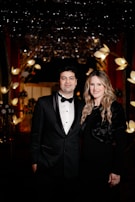 A formally dressed couple stands closely together in an elegant setting. Behind them, the background is adorned with decorative lights and floral arrangements, creating a luxurious atmosphere.