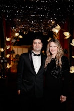 A formally dressed couple stands closely together in an elegant setting. Behind them, the background is adorned with decorative lights and floral arrangements, creating a luxurious atmosphere.