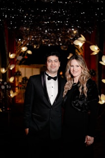 A formally dressed couple stands closely together in an elegant setting. Behind them, the background is adorned with decorative lights and floral arrangements, creating a luxurious atmosphere.