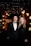 A formally dressed couple stands closely together in an elegant setting. Behind them, the background is adorned with decorative lights and floral arrangements, creating a luxurious atmosphere.