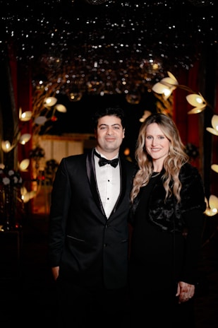 A formally dressed couple stands closely together in an elegant setting. Behind them, the background is adorned with decorative lights and floral arrangements, creating a luxurious atmosphere.