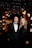 A formally dressed couple stands closely together in an elegant setting. Behind them, the background is adorned with decorative lights and floral arrangements, creating a luxurious atmosphere.