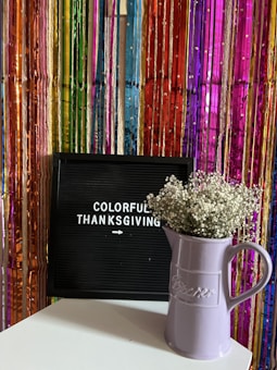 A vibrant backdrop made of colorful streamers in shades of pink, purple, red, blue, and green set the scene behind a black letter board. The board displays the words 'Colorful Thanksgiving' in white letters, with a small arrow pointing right. In front of the board, a light purple ceramic pitcher holds a cluster of small, delicate white flowers, possibly baby's breath, placed on a white surface.