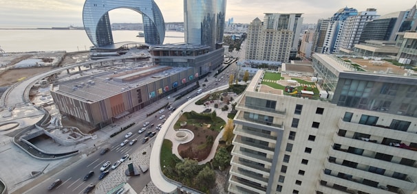 An urban landscape with high-rise buildings, roads filled with vehicles, and a distinctive modern architectural structure featuring a large arch. The scene includes a waterfront area with a view of the sea and a mix of contemporary and classic architectural styles. Green spaces and rooftops with gardens or terraces add to the city's aesthetic.
