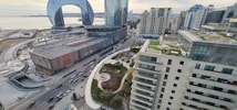 An urban landscape with high-rise buildings, roads filled with vehicles, and a distinctive modern architectural structure featuring a large arch. The scene includes a waterfront area with a view of the sea and a mix of contemporary and classic architectural styles. Green spaces and rooftops with gardens or terraces add to the city's aesthetic.