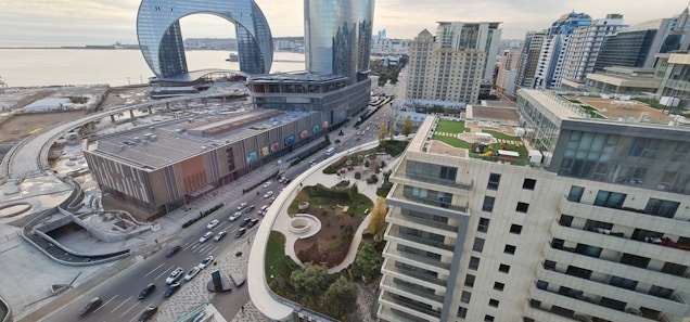 An urban landscape with high-rise buildings, roads filled with vehicles, and a distinctive modern architectural structure featuring a large arch. The scene includes a waterfront area with a view of the sea and a mix of contemporary and classic architectural styles. Green spaces and rooftops with gardens or terraces add to the city's aesthetic.