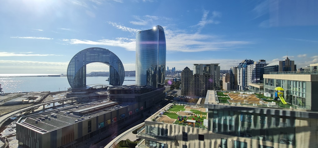 A scenic view of a modern cityscape with distinctive architecture, including a building with a circular opening and a sleek, glass skyscraper. The scene overlooks a body of water, reflecting sunlight, with a coastline visible in the background. Rooftops are adorned with green spaces and play areas.