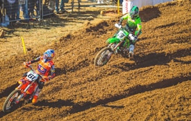 Two motocross riders compete on a dirt track, showcasing vibrant gear and motorcycles. The rider on the left is wearing a colorful helmet and orange attire riding an orange bike, while the rider on the right is in green gear on a green bike. The background is fenced, with spectators observing the race.