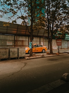 A yellow taxi is parked by the roadside next to a row of trash bins. The background features corrugated metal fencing and a few trees with autumn leaves. Signage in the vicinity suggests a commercial area.