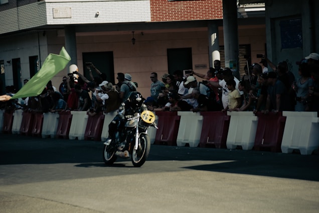 A motorcyclist wearing a helmet speeds on a road lined with red and white barriers. A crowd of spectators watches excitedly, many capturing the moment with their smartphones and cameras. A person in the foreground is waving a large green flag.