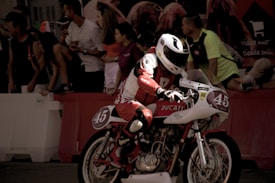 A motorcyclist in racing gear rides a classic Ducati motorcycle with number 45. The background includes a crowd of spectators, some of whom are sitting or standing on elevated surfaces, observing the event. The lighting gives the scene a dramatic and vintage feel.