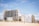 A row of beachfront high-rise buildings stands adjacent to a sandy beach, under a clear blue sky with light clouds. Some small vegetation grows on the dunes just before the buildings.