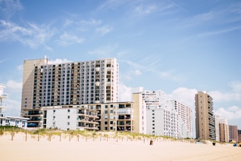 A row of beachfront high-rise buildings stands adjacent to a sandy beach, under a clear blue sky with light clouds. Some small vegetation grows on the dunes just before the buildings.