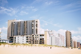 A row of beachfront high-rise buildings stands adjacent to a sandy beach, under a clear blue sky with light clouds. Some small vegetation grows on the dunes just before the buildings.