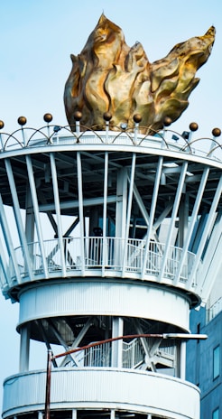 A large ornamental structure topped with a stylized golden flame. The base of the structure is composed of white metal beams and rings, resembling a tower. The flame is detailed with swirling patterns and sits atop a platform supported by vertical beams.