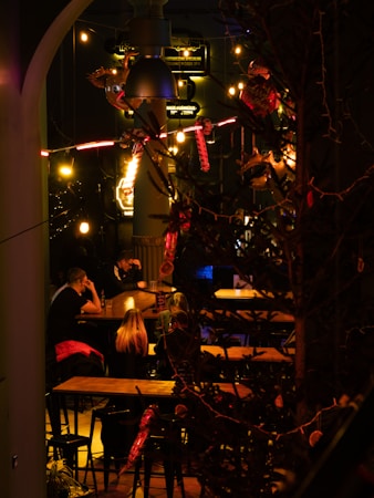 An indoor setting with a cozy, dimly lit atmosphere, featuring decorative lights and a couple of tables with people sitting. The foreground includes branches and decorative elements, while vintage-style signs and hanging bulbs create an ambient feel.