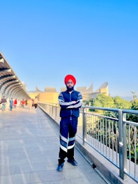 A person wearing a red turban and a blue tracksuit stands on a walkway bordered by metallic railings. The clear blue sky stretches above, while an architectural building with unique shapes is visible in the background. Several people are present further down the walkway, some dressed in colorful attire, walking in both directions.